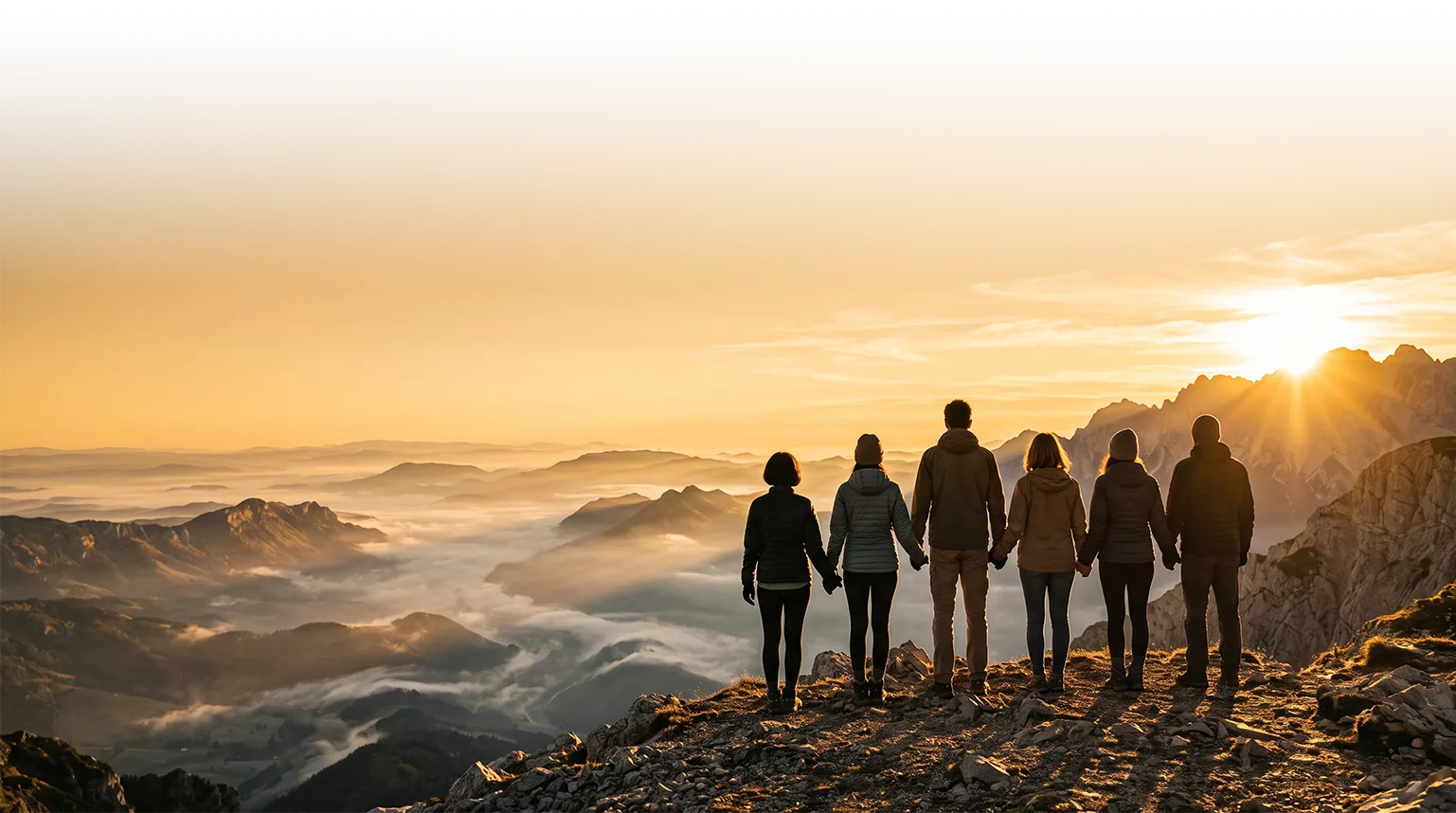 Six people holding hands on a mountain ridge, watching the sun rise over a sea of clouds.